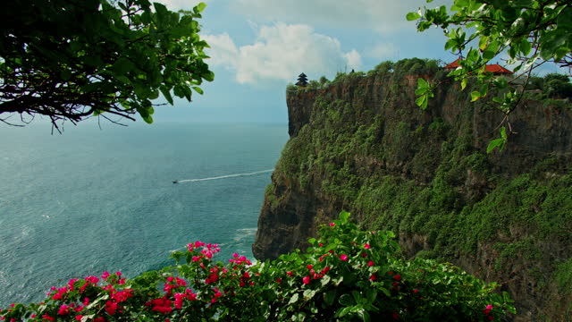 Natural landscape Pura Luhur Uluwatu temple in Bali Island Indonesia. Beautiful huge cliffs with exotic plants, red magenta flowers against blue Indian Ocean. Famous historic balinese monument.