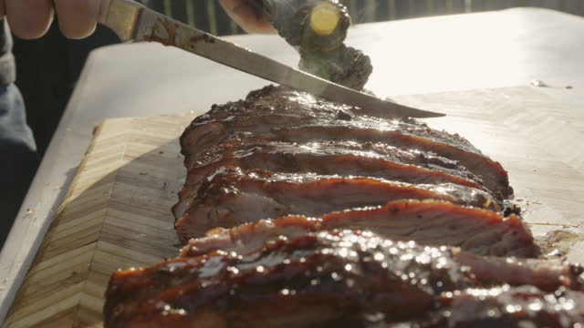Slow-motion shot of a rack of Barbecue pork spare ribs on a rustic wooden cutting board being sliced in preparation for a barbecue dinner