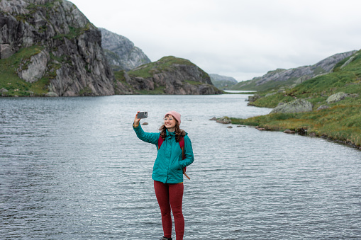 Young Caucasian woman standing near the lake and making selfie on  the background of mountains