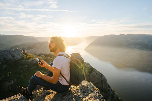 Young Caucasian man sitting on Preikestolen and using smartphone