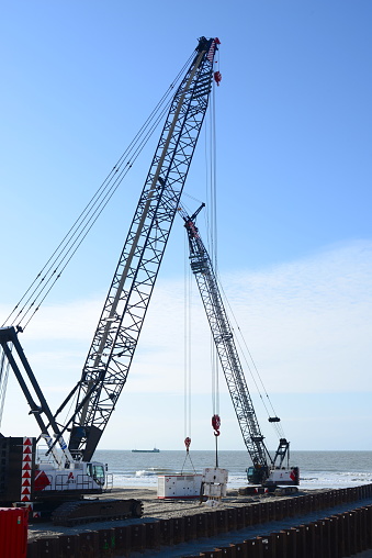Blankenberge, West-Flanders, Belgium - April 26, 2023: Cobelco construction crane with a bucket to grab the corrugated sheet profiles out from the sand and the other crane holding on cables an energy battery power cabin connected to the bucket grab