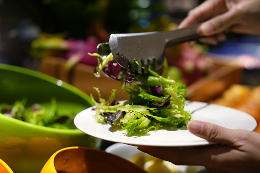 Assorted vegetables in bowls at the salad bar, cropped image of a woman is at the salad bar, picking up fresh salad with serving tongs.