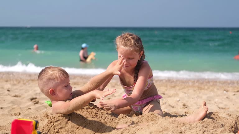 A sister buries her brother on the beach in the sand. Children in the summer at the sea play a girl and a little boy. Summer time. Travel.