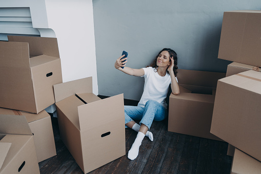 Home owner is taking selfie in new luxurious apartment. Happy woman unpacking cardboard boxes. Girl is taking photo on phone and sitting on the floor. Independence and success concept.