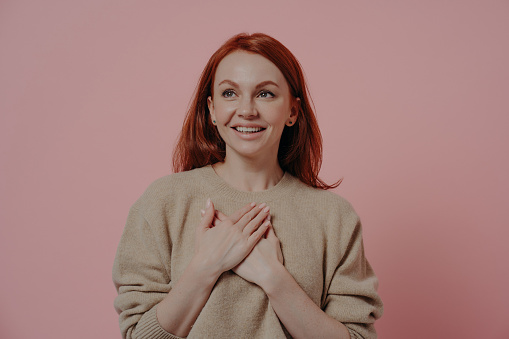 Isolated studio shot of young happy red-haired woman putting hands on chest, touching heart, looking aside with gratitude and thankfulness while standing in beige jumper isolated over pink background