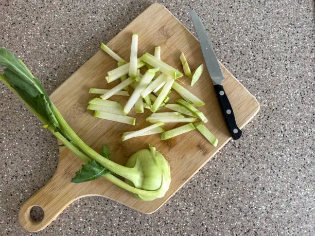Sliced ​​kohlrabi cabbage, half with leaves and next to it cut into strips, knife, on a wooden cutting board on a marble chip kitchen worktop, top view