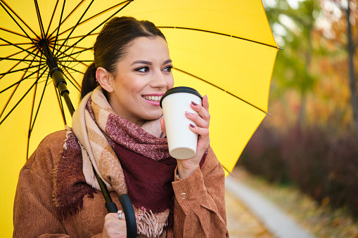 Caucasian young woman with coffee and yellow umbrella smiling in autumn.