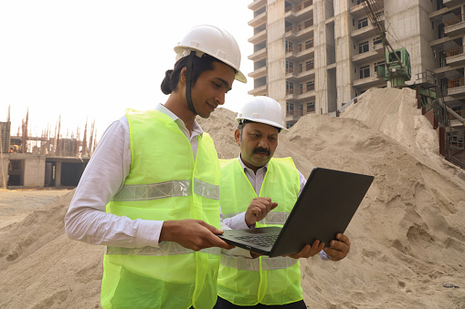 Happy Indian construction engineer working on a construction site reviewing and analysing the work in progress. Manager examining or inspecting the work flow and the quality of the work with engineer. They are holding the laptop and working.