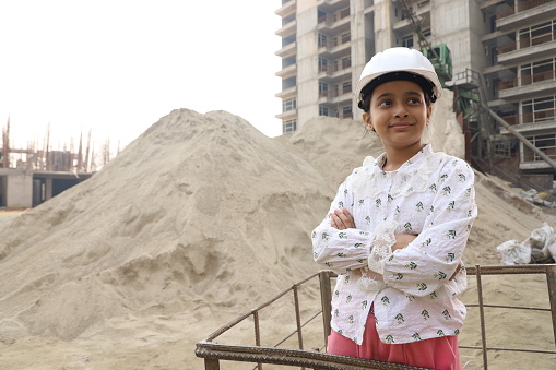 Teenage Indian girl acting as a construction engineer manager working on a construction site with the team and reviewing and analysing the work in progress. The professional girl holding the blueprint roll in her hand. Cheering up on job. Happy to take responsibilities.