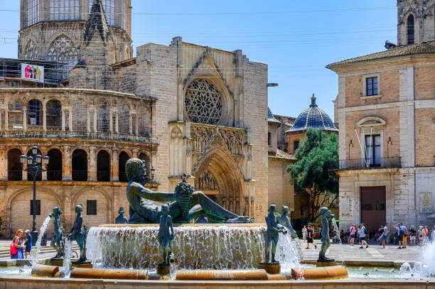Valencia, Spain - April 8, 2023: View of the historic Plaza de la Virgen featuring the striking Turia Fountain and Cathedral in the foreground, surrounded by other buildings. Incidental people are standing and walking in the background under a clear blue sky.