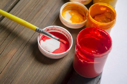 Red paint in a jar on a wooden table. The brush lies on the hook from the jar of paint. Bright colors on the table. Gouache is red.