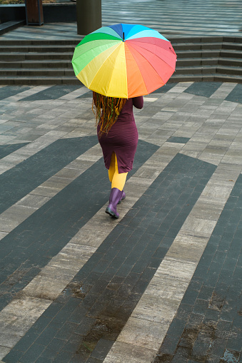 Rainbow umbrella on city street . Walking on a rainy spring day. Woman wearing rubber boots following the line. Bright colors