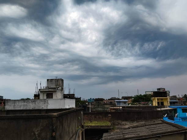 This photograph captures the dramatic scene of a cloudy afternoon before a heavy rainfall in old Kolkata city. The dark, billowing clouds are shown forming intricate designs in the sky, promising relief from the scorching summer. The image showcases the bustling city life in the background, waiting for the rain to arrive.