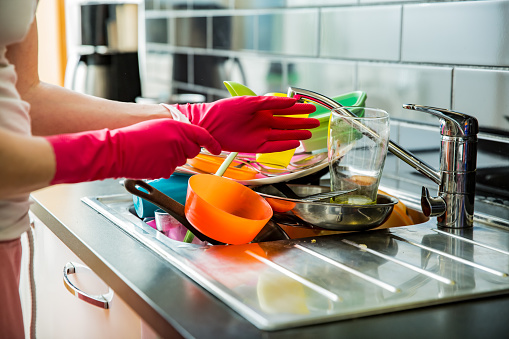 Kitchen sink full of dirty dishes. Woman in pink rubber gloves doing the dishes, using brush. Dark colored countertop in modern kitchen. Household chores concept