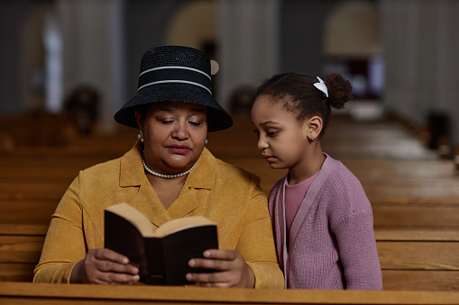 Grandmother reading Bible to her granddaughter while they sitting on bench in church