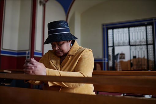 Elegant woman in hat sitting on bench with her eyes closed and praying in church