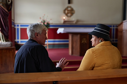Rear view of senior priest discussing the ceremony with woman while they sitting on bench during her visit to church