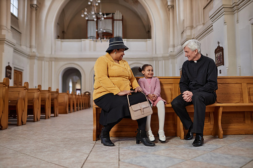 African American grandmother talking with priest together with her little granddaughter while they sitting on bench in church