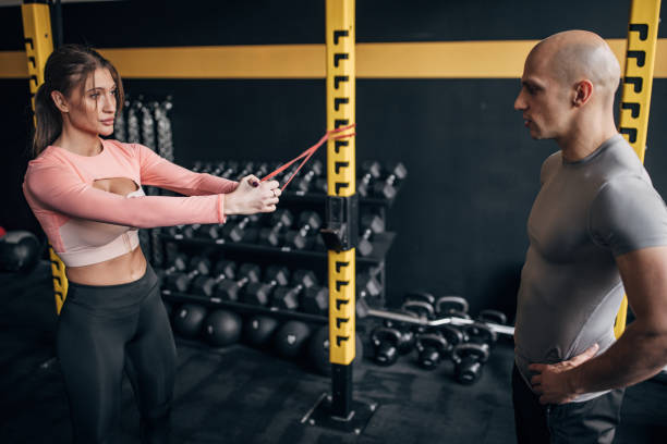 Woman with fitness trainer Two people, fit female training with a rubber strap, while his fitness trainer is assisting her in gym. personal trainer cost stock pictures, royalty-free photos & images