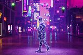 Metaverse Cyberpunk Style City With Artificial Man Walking On Street, Neon Lighting On Building Exteriors, Flying Cars And Drones