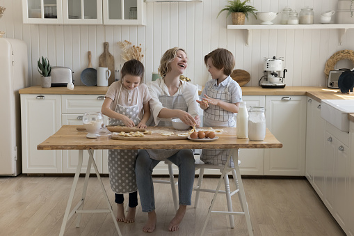 Cheerful mother and two cute little kids wearing aprons, baking dessert in home kitchen, preparing delicious bakery food together, rolling, shaping dough, talking, chatting, laughing