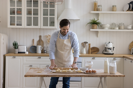 Positive handsome baker man wearing apron rolling dough on floury table, baking in home kitchen interior, enjoying craft bakery food preparation, cooking, culinary hobby, smiling