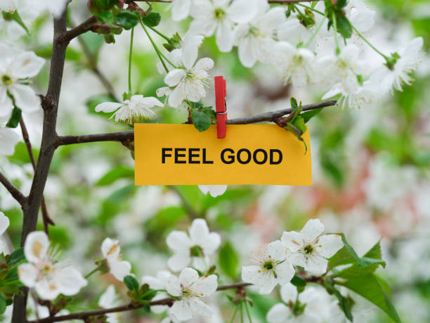 A yellow paper note with the phrase Feel Good on it attached to a branch with cherry blossoms on it with a clothes pin