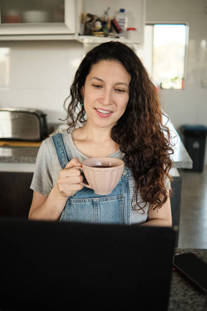 scatto verticale di una donna latina davanti al suo laptop sorridente con una tazza di caffè tra le mani che lavora da casa - beautiful looking at camera studio shot vertical foto e immagini stock