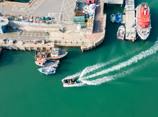 Aerial drone view of a speed boat coming into a harbour. Boats are tied and moored beside tie the concrete pier and dock area. The engine and speed of the boat is causing stern wake in the water behind the boat.