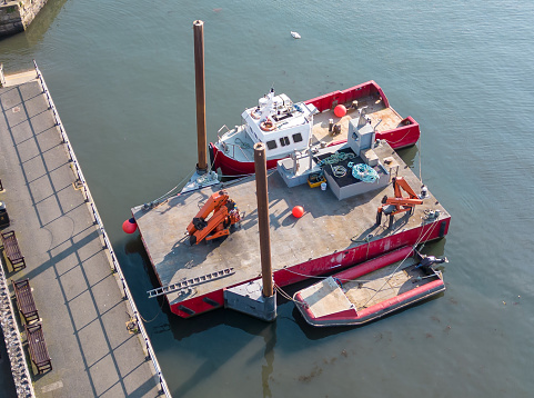 Aerial drone view of a small pontoon floating in water, with two cranes fixed to it and two small boats moored beside it in the harbour of a Devon fishing village.