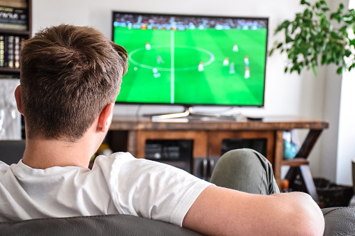 Young man sitting on a couch at home watching soccer game on TV