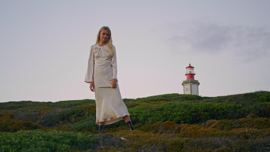 Blonde model posing nature green hill. Cute woman watching camera taking different positions at lighthouse background. Calm girl in naked back dress standing sunset environment. Tender lady alone