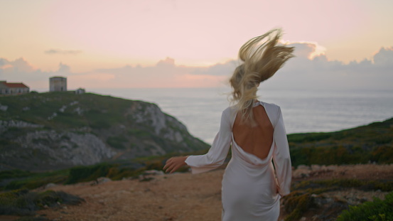 Blonde woman moving castle seashore back view. Happy girl running beautiful empty valley. Carefree lady wearing dress having fun on nature alone. Attractive model looking over shoulder at camera