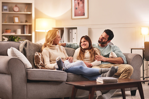 Relax, mom and dad with teenager on sofa, conversation and quality time with smile at family home in evening. Father, mother and daughter on couch, talking and fun together with bonding in happiness.