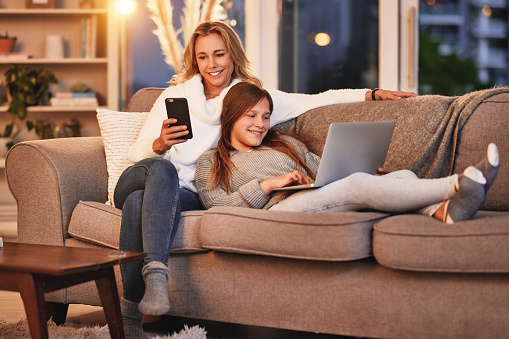 Mother, teenager and sofa with laptop, phone and relax with quality time online in evening at home. Lounge, happy woman and daughter on couch, browsing internet or social media together in happiness.