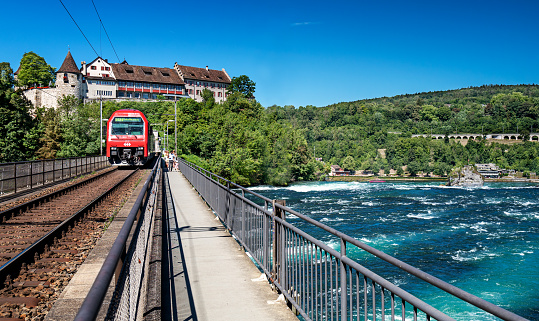 Zurich, Switzerland - July 11, 2022: The SSB train traveling over the Rhine River near the Rhine Falls with the Schloss Laufen Castle in the background.