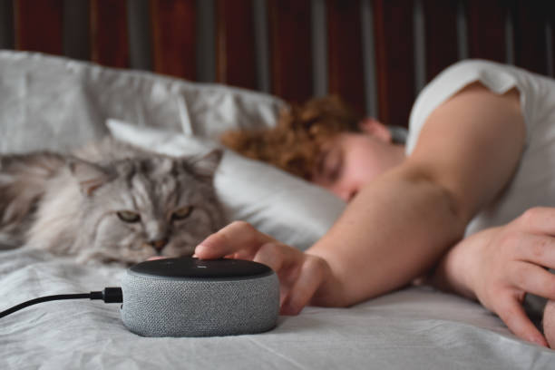 Young man sleeping with a cat and pressing snooz button on virtual assistant