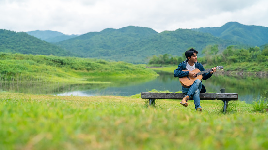 Young Asian man relax and enjoy outdoor lifestyle travel nature camping in forest mountain on summer holiday vacation. Handsome guy drinking coffee and playing guitar by the lake in the morning.