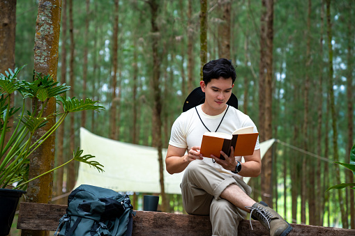 Asian man relax and enjoy outdoor lifestyle camping in forest mountain on summer holiday travel vacation. Handsome guy resting on outdoor chair and reading a book in pine tree forest in the morning.