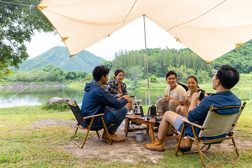 Group of Happy Asian Man and woman friends having breakfast and making brewed coffee at camp in the morning. People enjoy and fun outdoor lifestyle travel nature and camping together on summer vacation.