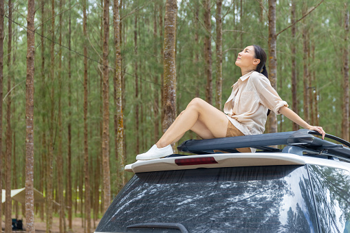 Asian woman enjoy and fun outdoor lifestyle road trip travel nature and camping in forest mountain on summer holiday vacation. Attractive girl sitting on car roof rack by the road with looking pine tree forest.