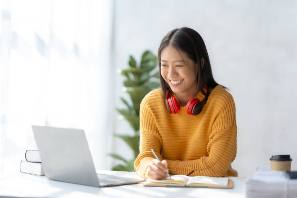 istock Asian businesswoman sitting taking notes and using laptop computer at home happily working. Young Asian female student taking notes and using laptop in library. 1486660757