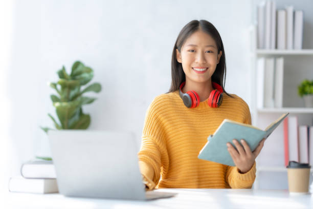 istock Asian businesswoman sitting taking notes and using laptop computer at home happily working. Young Asian female student taking notes and using laptop in library. 1486660774