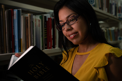 In the evening Latino woman readers in the library read a book between the shelves in the dim evening light. She read the new Bible. She is posing with a book, a portrait, looking at the camera.