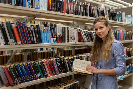 A white jung woman reader is reading a book in the dim evening light between the shelves in the library. Portrait, looking at the camera.