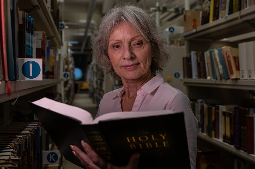 A older white woman reader is reading a book in the dim evening light between the shelves in the library.  She reads the bible.