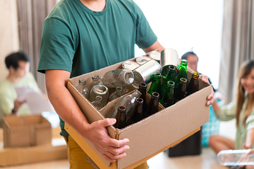 Asian man bends down to pick up and hold onto a big box of mixed materials to be reused or recycled while looking at camera as his other five Asian friends continue to sorting between paper, glass bottles, plastic bottles and cardboard boxes indoors on the ground of the living room.