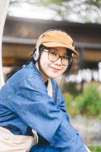 People mental therapy with at outdoor nature lifestyle concept. Young asian woman relax listening music with fashion headphone wearing hat denim jacket and eyeglasses. Vertical portrait.