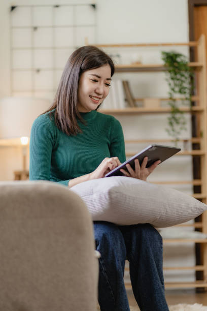 istock Happy asian girl holding tablet device using digital tablet technology sitting on sofa at home smiling woman using app online shopping 1486703315