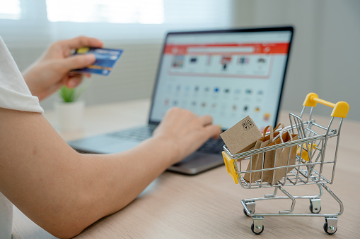 Online shopping-boxes or parcels are placed on the table and shopping carts. Blurred background, woman use credit card to make online purchases on laptop. Online service. New normal shopping delivery.